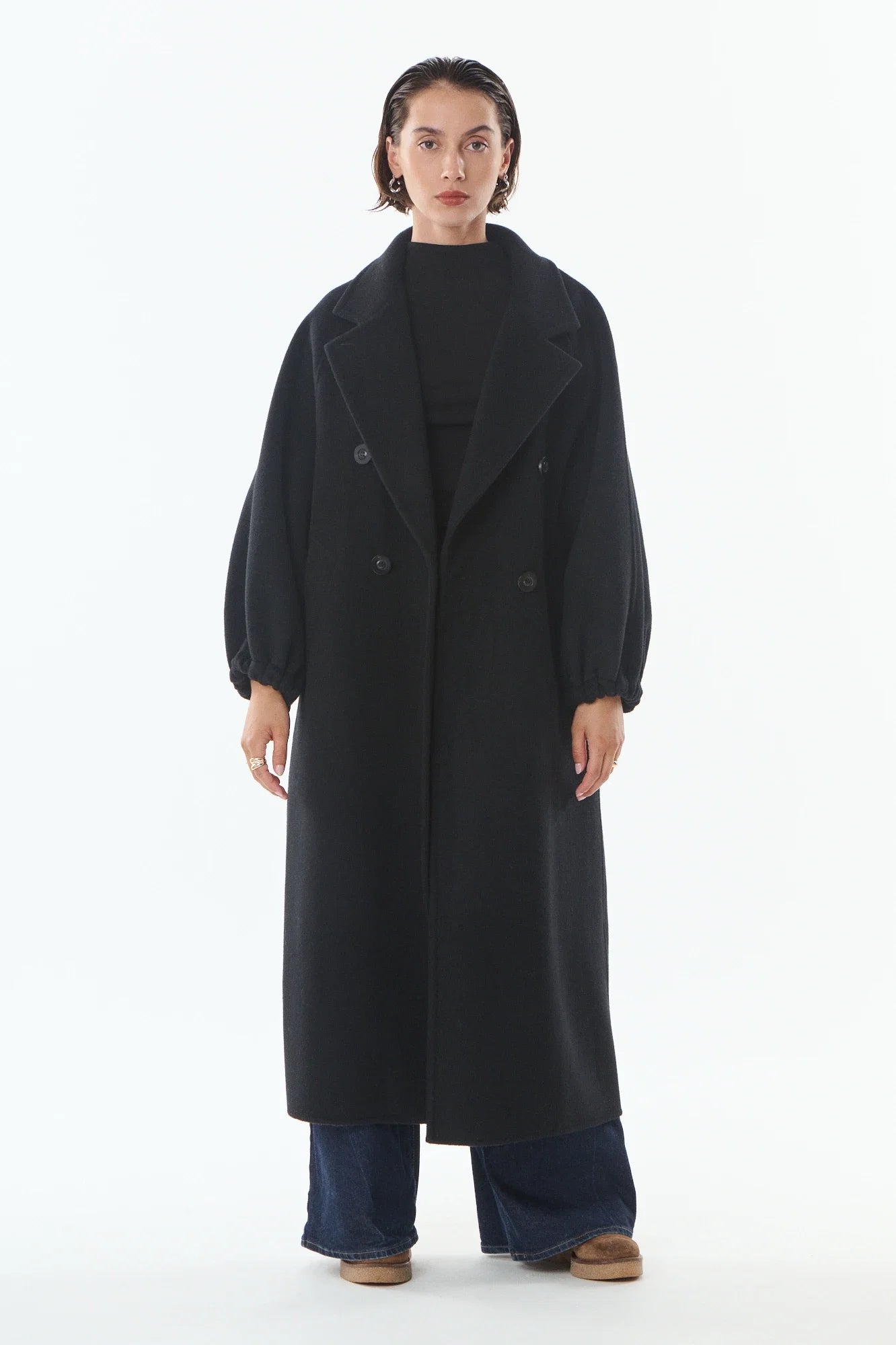 A model stands against a plain white studio background wearing a long black double-breasted coat over a black top, wide-leg blue jeans, and brown shoes.
