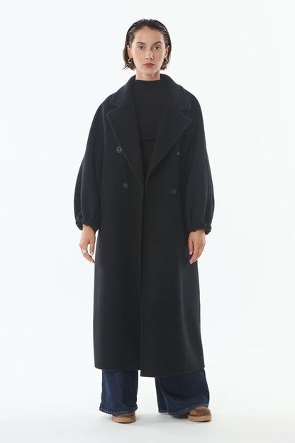 A model stands against a plain white studio background wearing a long black double-breasted coat over a black top, wide-leg blue jeans, and brown shoes.