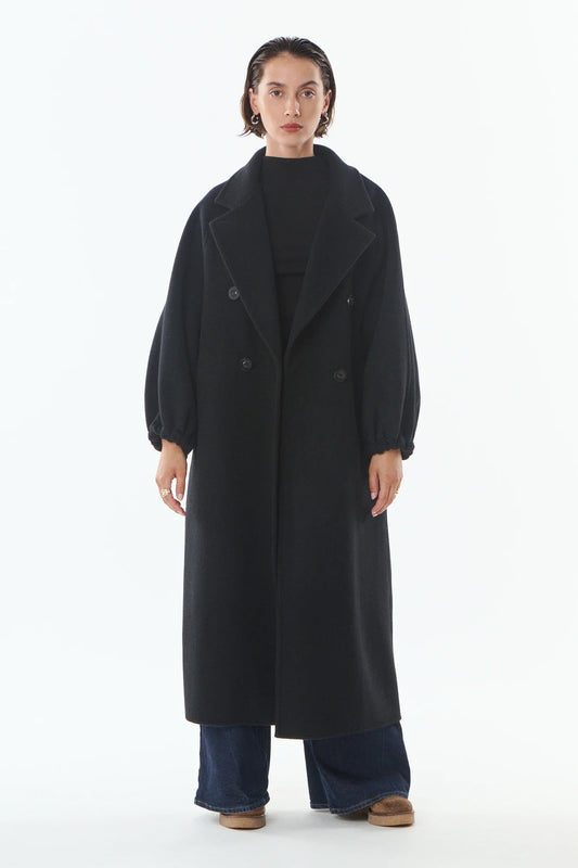 A model stands against a plain white studio background wearing a long black double-breasted coat over a black top, wide-leg blue jeans, and brown shoes.